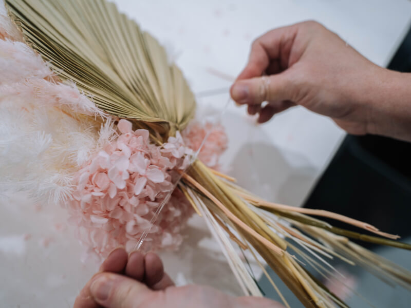 Hands tying up a dried flower arrangement with pink petals and a dried palm leaf 